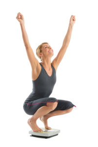 Happy Woman With Arms Raised Crouching On Weight Scale